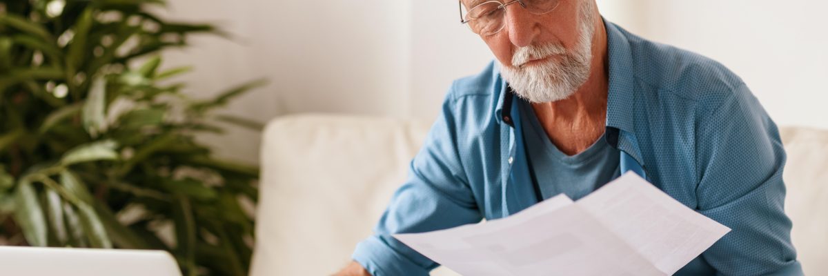 Elderly serious man in glasses reading document and making notes in laptop while sitting at table and working on freelance project at home